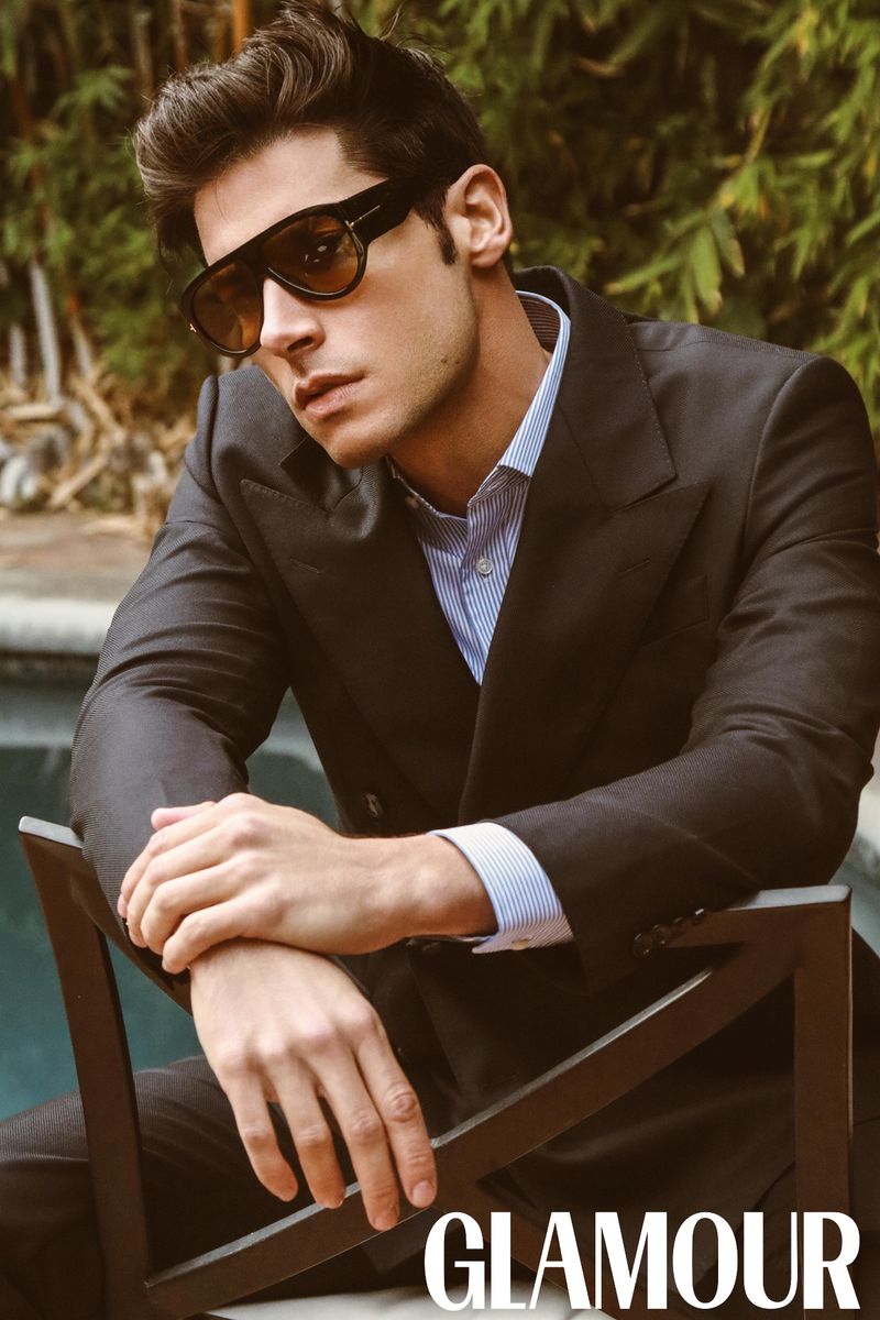 Portrait of a male model wearing a dark suit and sunglasses, seated on a chair with foliage in the background, magazine cover.