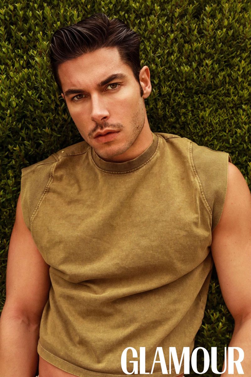 Portrait of a brunette male model with short hair, wearing a tan t-shirt, posing against a dense green foliage background, with Glamour logo.