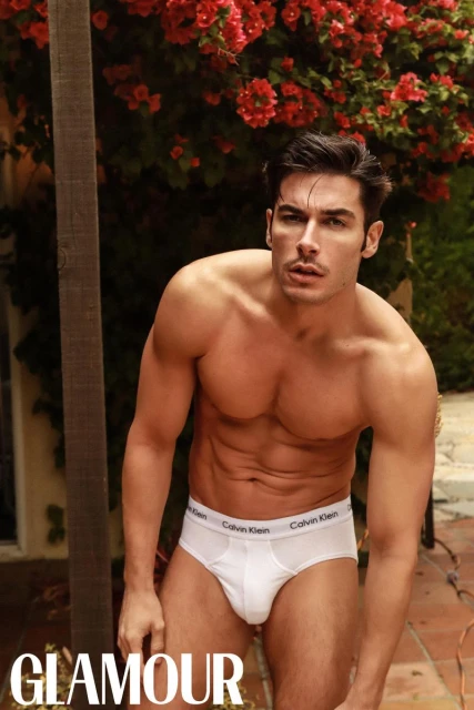 Andrea Denver posing in white briefs outdoors against blooming red bougainvillea flowers.