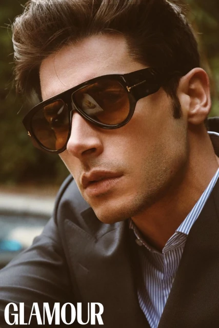Close-up portrait of model Andrea Denver wearing oversized black sunglasses, a dark blazer, and a striped dress shirt.