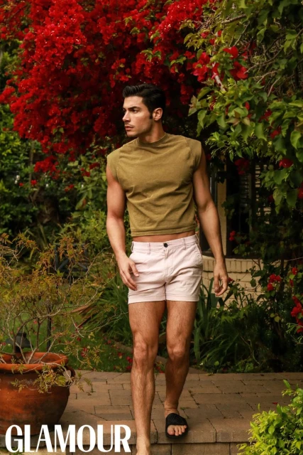 Model Andrea Denver walking outdoors past vibrant red flowers wearing a green sleeveless top and light shorts.