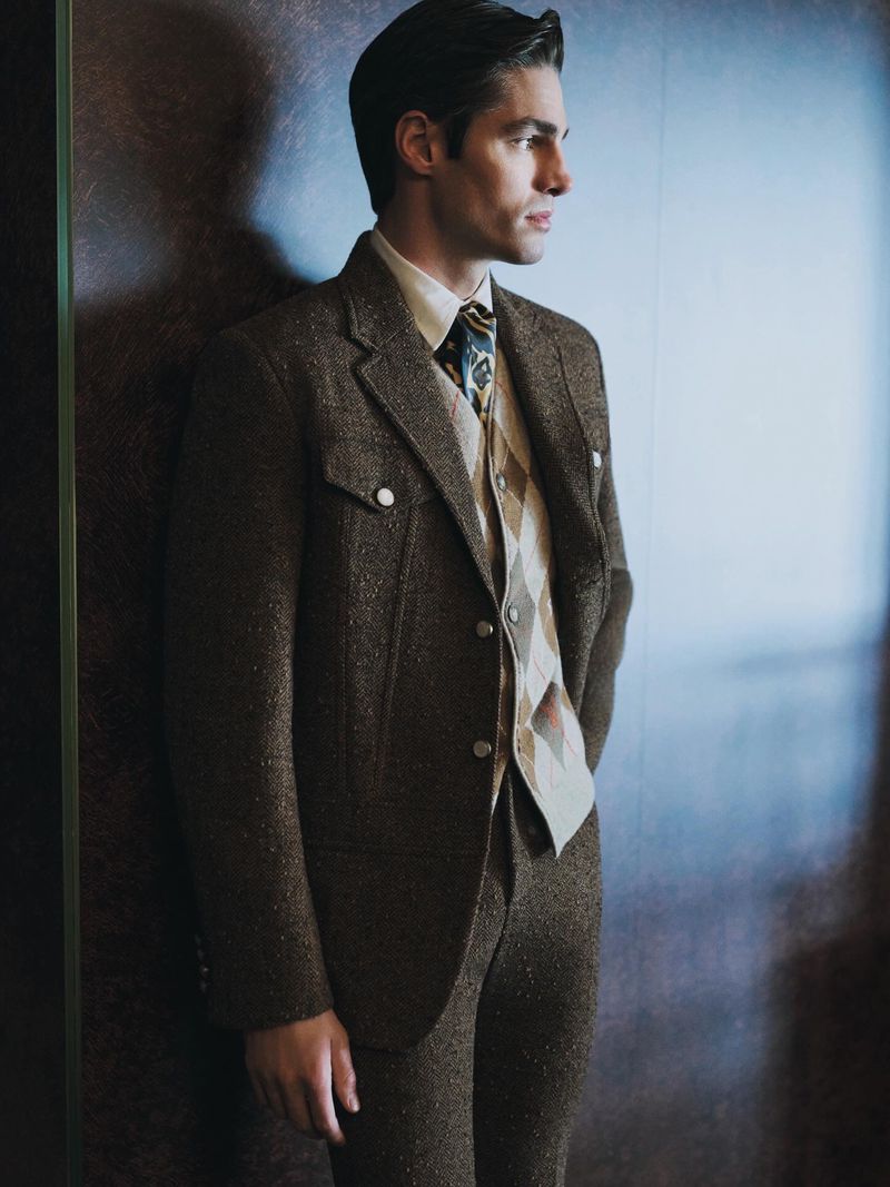 A man in a brown tweed suit poses indoors with a patterned tie and vest, standing against a wall with soft lighting. He is looking away.