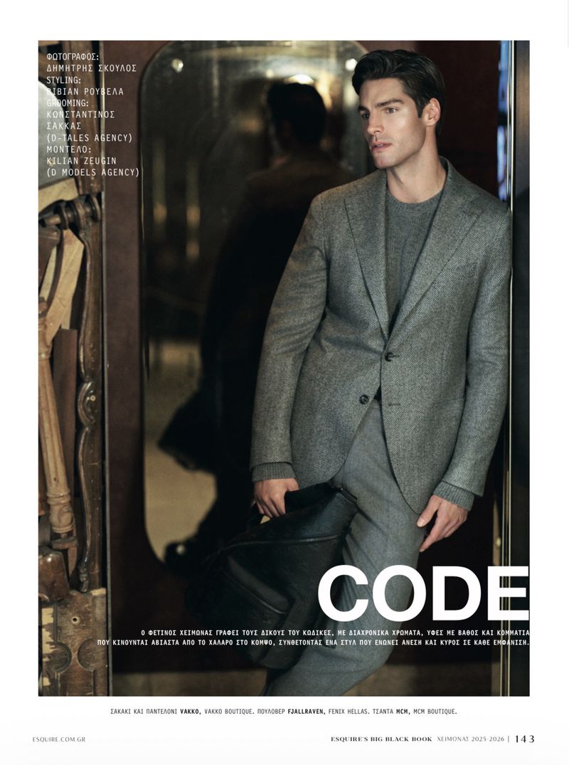 Male model poses in a gray suit and sweater, holding a black bag indoors with ornate decor. Fashion editorial shot.