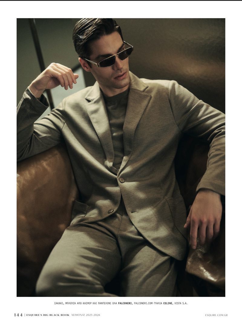 A male fashion model in a beige suit and sunglasses reclines on a leather chair in a studio with soft, flattering light.
