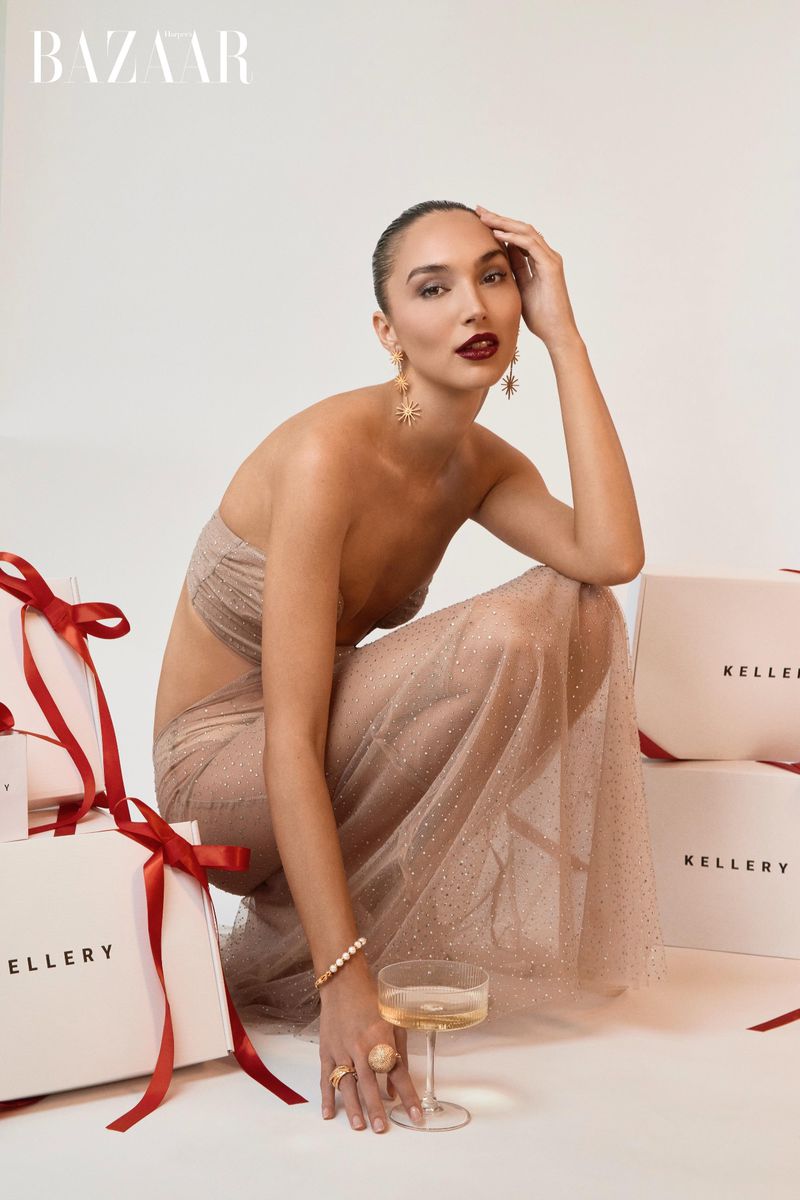 Elegant woman in a sheer dress poses with Kellerry shopping bags and champagne against a white background for a beauty ad.