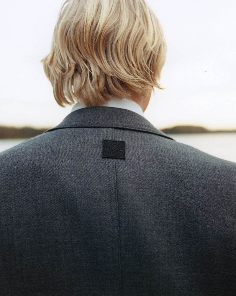 Back view of a light-haired adult wearing a gray blazer and white shirt standing outdoors with a body of water in the background.