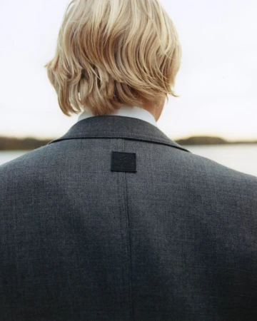 Back view of a light-haired adult wearing a gray blazer and white shirt standing outdoors with a body of water in the background.