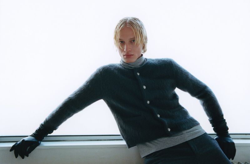 Young adult with blond hair, wearing a gray turtleneck and dark fuzzy cardigan, leans on a white windowsill in a fashion editorial.