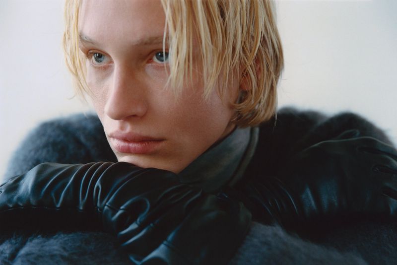 Close-up of a fair-skinned adult male with blonde hair, wearing black leather gloves and a fuzzy gray sweater, posing indoors.
