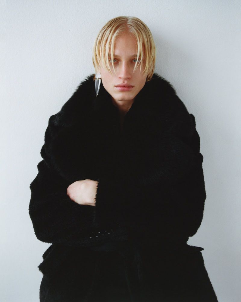Androgynous fashion portrait of a person with blond hair in a black fur coat against a pale white wall, gazing at the camera.