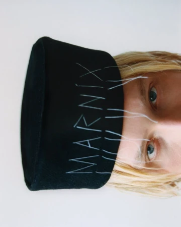 Close-up of a fair-skinned adult wearing a black beanie with the brand name 'Marnix' in white, set against a plain white background.