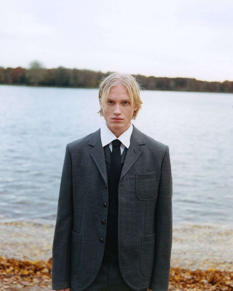 A blond-haired male model in a grey suit stands near a lake under a cloudy sky, creating a somber, fashion-editorial style portrait.