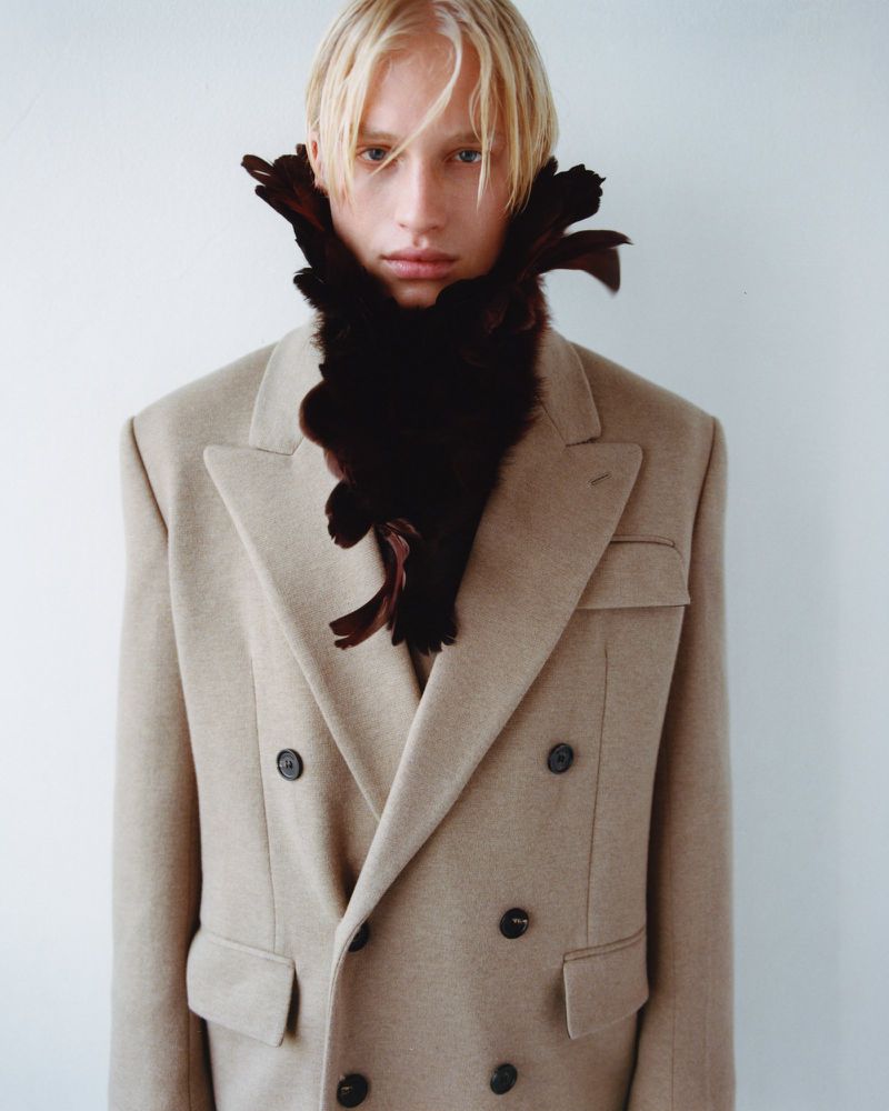 A fair-skinned male fashion model with platinum blonde hair poses in a light beige wool suit and a brown feather boa in a photo studio.