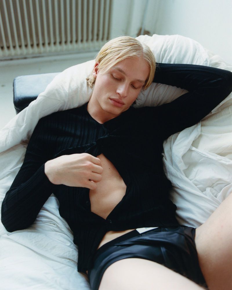 A blonde male model reclines on a white bed, wearing a black cardigan and black shorts, with natural light and a minimalist backdrop.