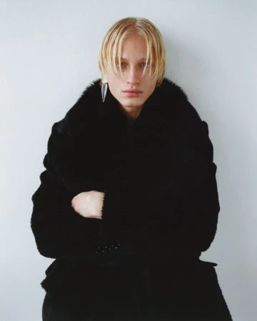 Blonde male model in an oversized black fur coat and pointed silver drop earring against a white studio background.