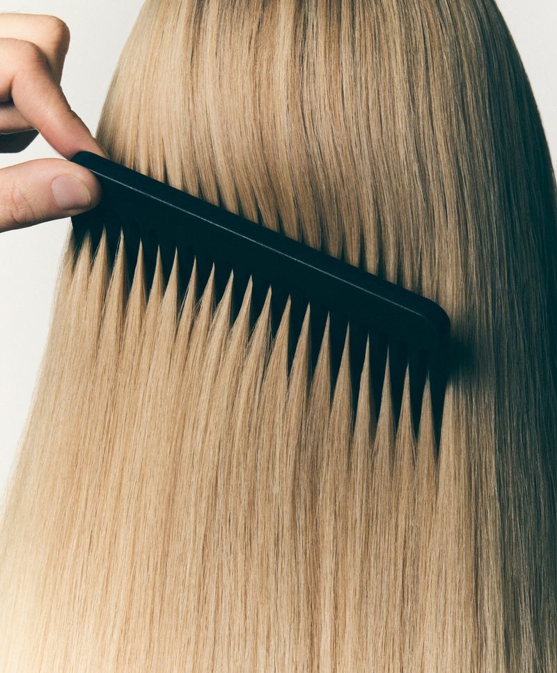 Close-up of a hand using a black comb on long, straight, blonde hair in a bright, neutral studio setting for a hair or beauty concept.