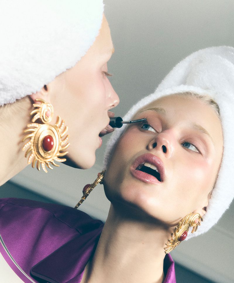 Close-up of a fair-skinned young woman applying mascara in the mirror with a white towel wrapped around her head; wearing gold jewelry.