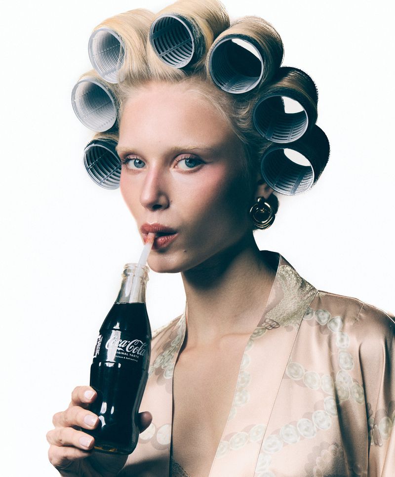 Adult female with hair rollers drinking Coca-Cola through a straw, wearing a silk robe against a white background in a beauty portrait.