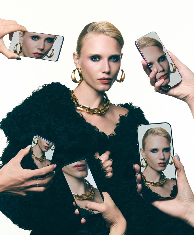 Fashion photo of a blonde woman with gold jewelry surrounded by hands holding phones, reflecting her face and style, against white.