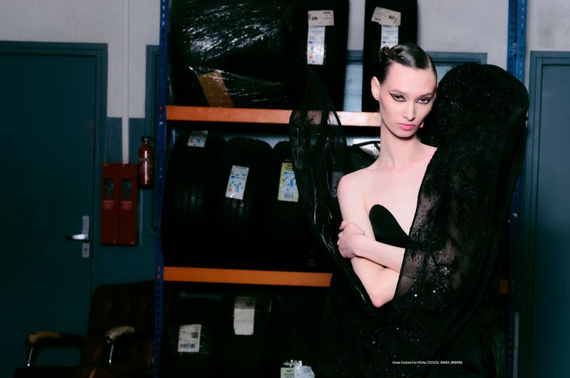 A model in a black dress poses in an industrial setting with tires stacked on shelves behind her, creating a fashion-editorial contrast.