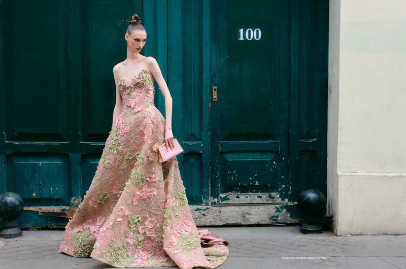 A woman in an elaborate formal gown poses in front of a dark green door with a small pink purse, creating a fashion editorial image.