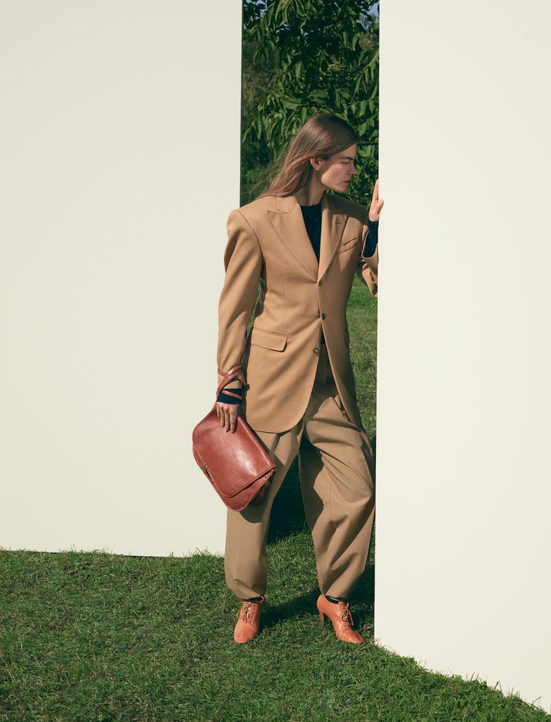Fashionable woman in tan suit with brown leather bag stands on green grass behind a modern white wall in an outdoor setting.