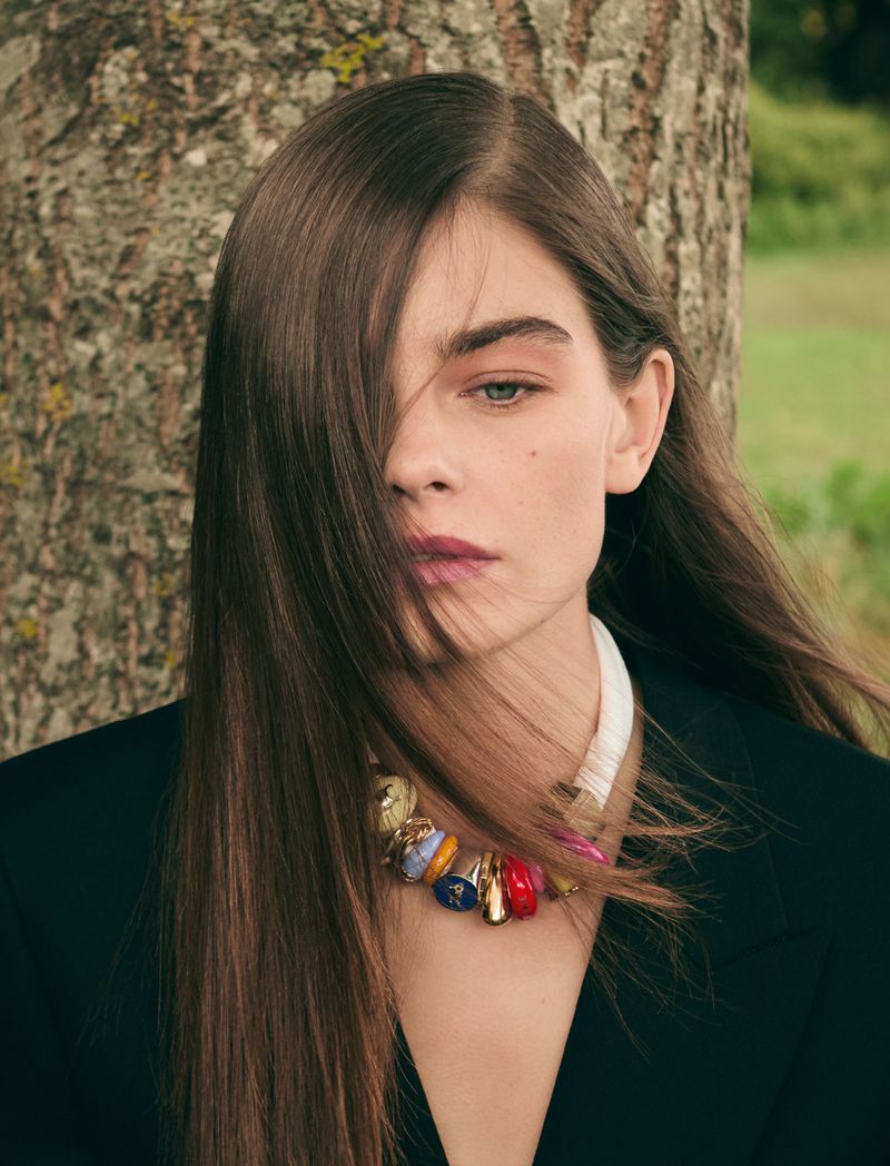 A woman with long brown hair poses outdoors wearing a black blazer and a colorful beaded necklace in a medium shot with a blurred background.