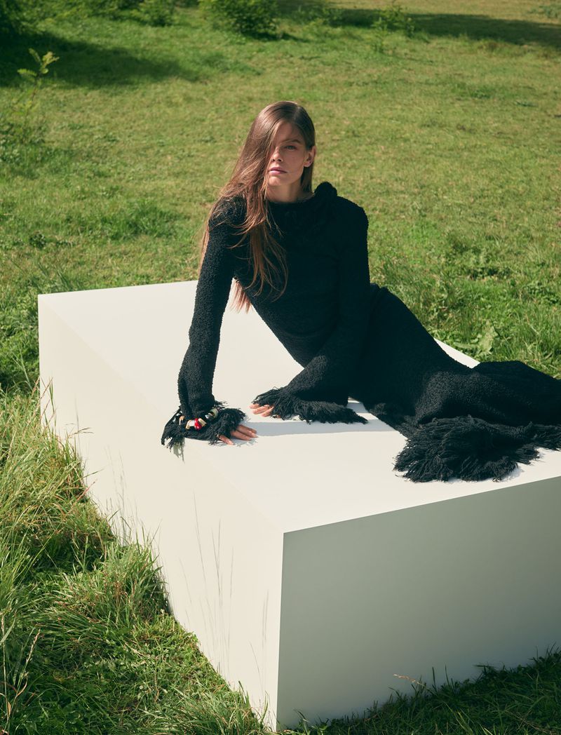 Woman in a black dress lying on a white block in a grassy field. The woman poses for a fashion shot in natural outdoor lighting.
