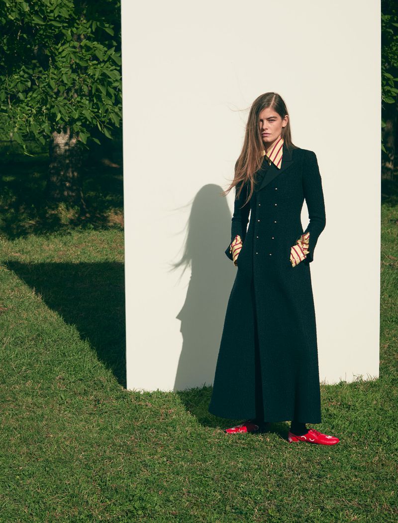 A woman in a long dark coat stands in a grassy field in front of a white wall, bathed in sunlight. She wears red shoes.