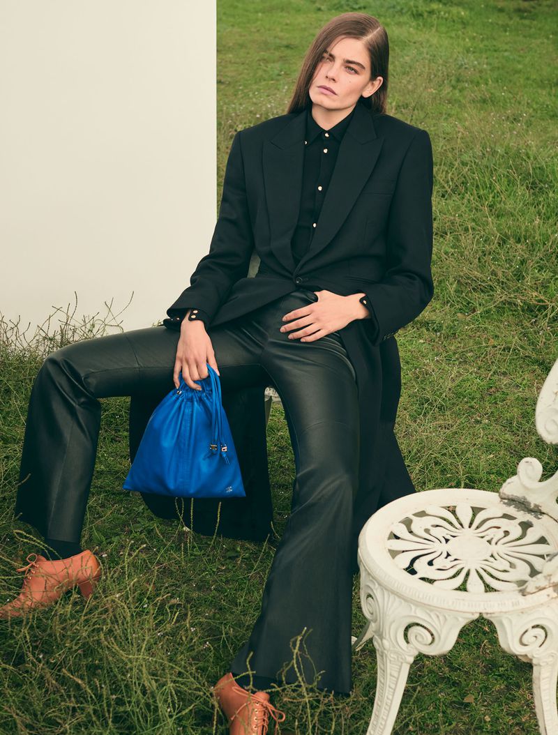 A model sits outdoors wearing a black suit, leather pants, and tan shoes, holding a blue bag, creating a fashion-editorial-style portrait.