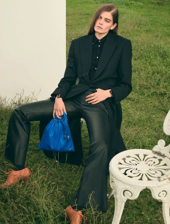 A model sits outdoors wearing a black suit, leather pants, and tan shoes, holding a blue bag, creating a fashion-editorial-style portrait.