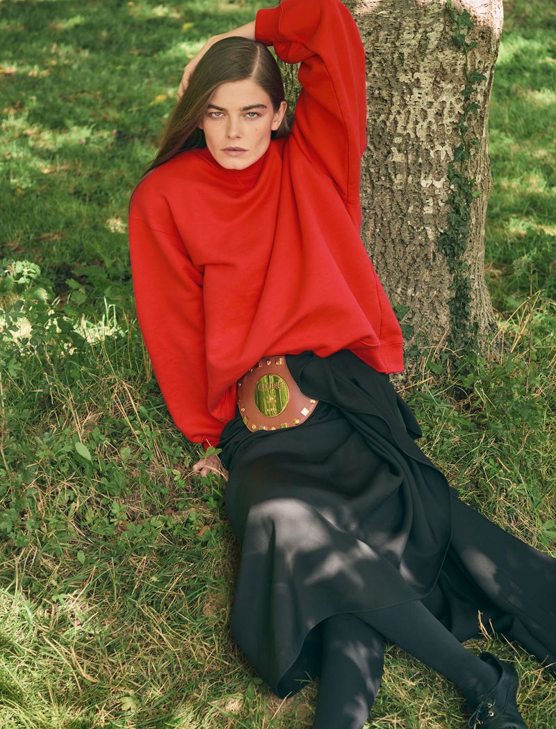 A woman in a red sweatshirt poses by a tree in a grassy field, showcasing fashion and outdoor lifestyle in natural lighting.