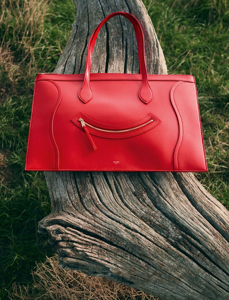 A red leather handbag sits atop a weathered tree trunk in a natural setting, highlighting its design and color against the organic backdrop.