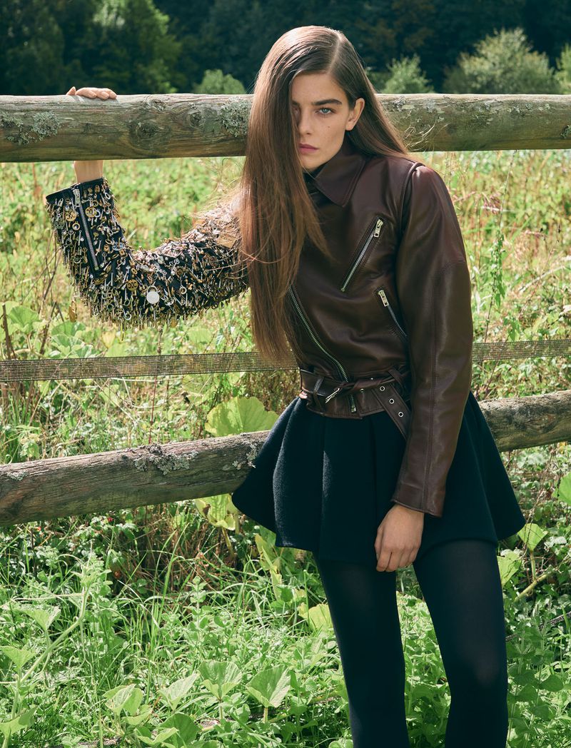 A woman poses against a wooden fence in a field, wearing a brown leather jacket, short black skirt, and dark tights. Fall fashion.