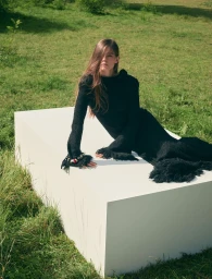 Merlijne Schorren reclining in a black fringed knit dress on a large white plinth in an open green grassy field.