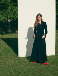 Model in long dark coat and striped shirt standing outdoors on grass before a tall white panel