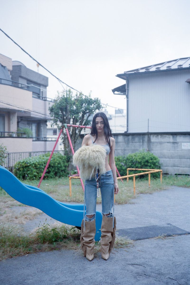 A woman in jeans and boots standing on a playground holding a fluffy bag, with a slide and apartment buildings in the background.