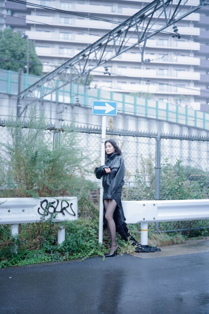 A woman in a black dress and coat stands on an urban street near a metal fence and greenery, with a building in the background.