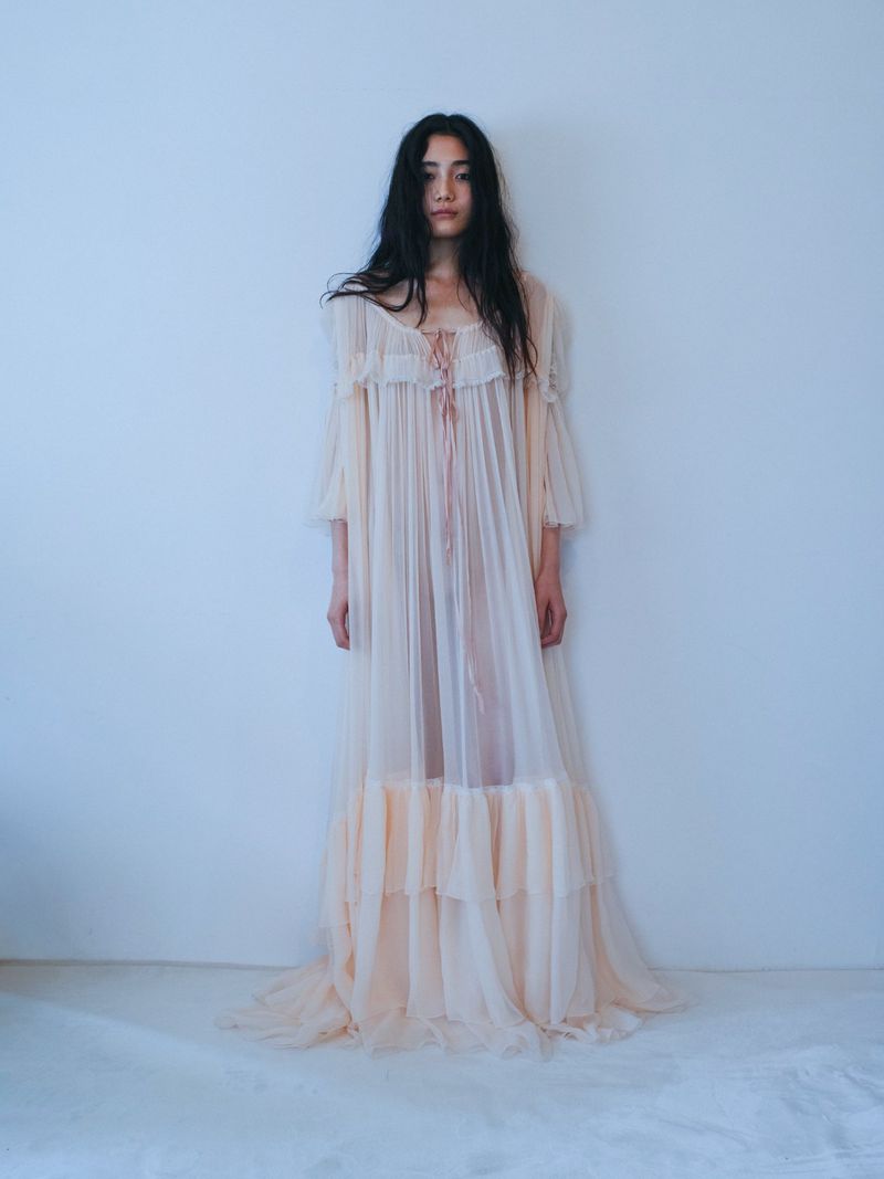 An adult female with dark hair is standing in front of a white wall, wearing a long, light-colored, semi-sheer dress with ruffled details.