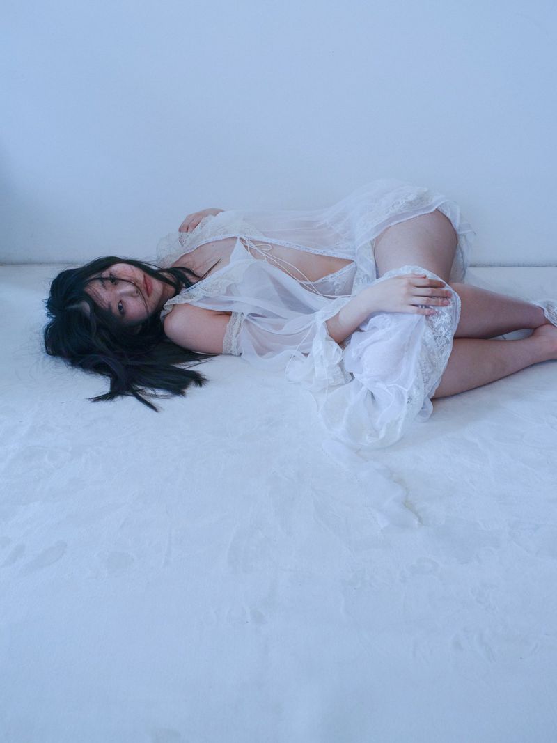 Woman in white lingerie lying on a white surface, creating a soft and sensual atmosphere with dark hair contrasting the bright surroundings.