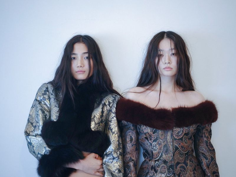 Two Asian women pose in embellished gowns with fur trim against a neutral wall, showcasing elegant and unique vintage fashion.