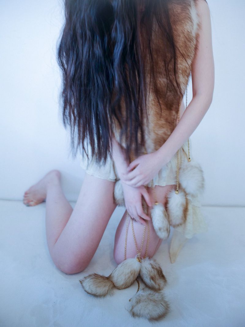 Seated woman with long dark hair wears a fur vest and decorative furry tails. Indoor setting with a minimalist aesthetic.