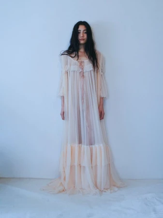 Model in a sheer pale pink ruffled long dress standing against a light grey studio wall for Carcy Magazine.