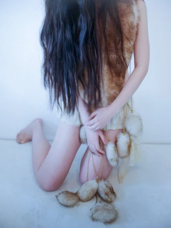Kneeling woman with long dark hair covering her torso, wearing a brown fur vest and cream skirt with hanging fur pom-poms.