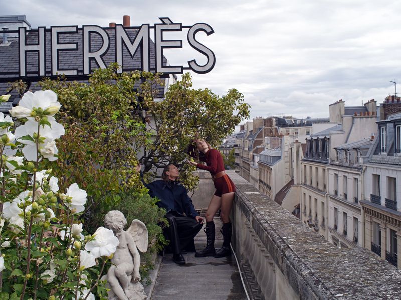 Fashion shoot on a rooftop featuring a man and a woman with the Hermès logo in the background and a cityscape in the distance.