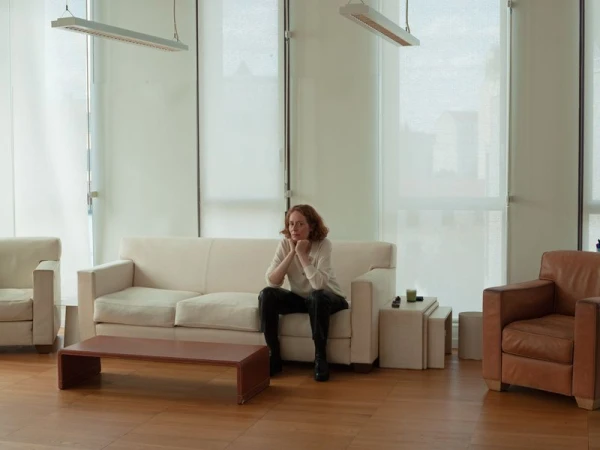 Woman sitting on a sofa in a room with large windows and light-colored furniture, wearing a white top and dark pants.