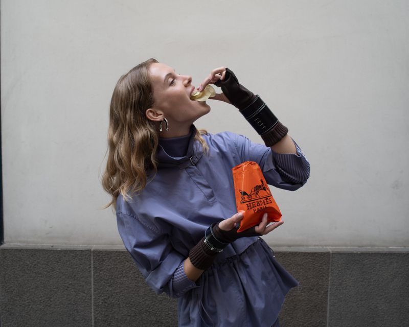 Woman in a periwinkle dress eating a snack out of an orange Hermes bag against a gray background with wrist warmers and hoop earrings.
