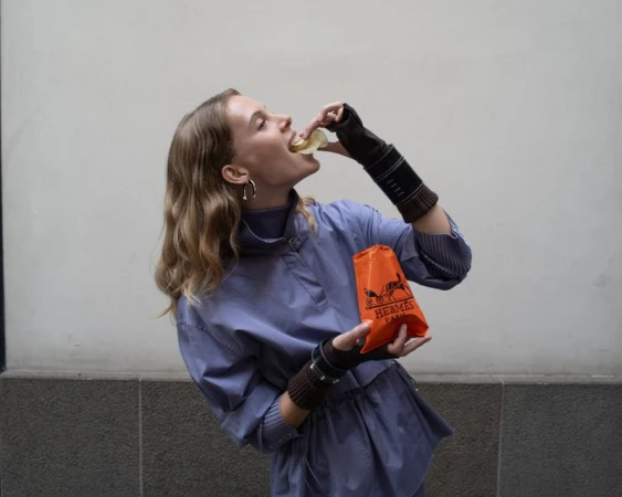 Woman in a periwinkle dress eating a snack out of an orange Hermes bag against a gray background with wrist warmers and hoop earrings.