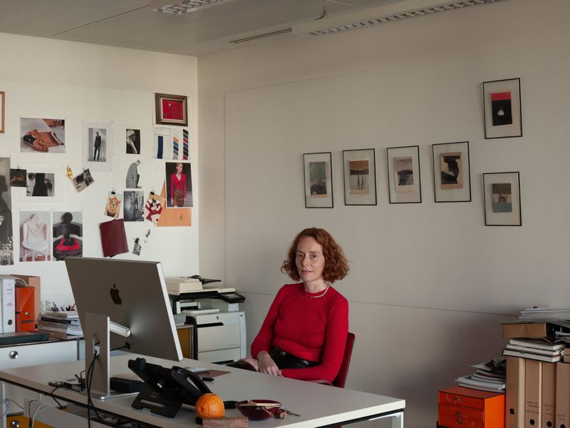 Adult woman sits at a desk in an office setting with a computer and artwork. The scene is brightly lit and captured in a medium shot.