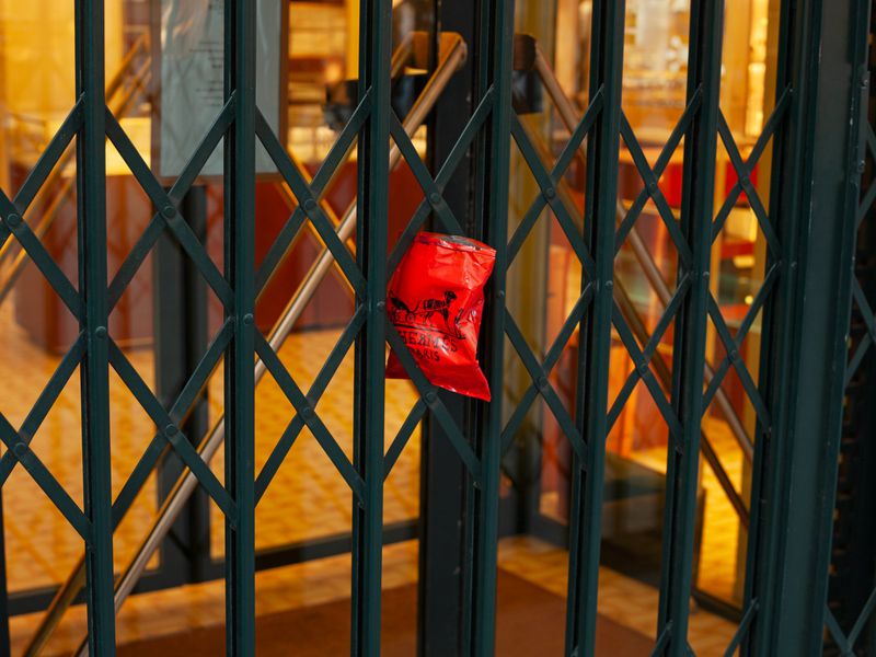 Green metal security gate of a store, with a red plastic bag hanging on it, with a blurred storefront in the background.
